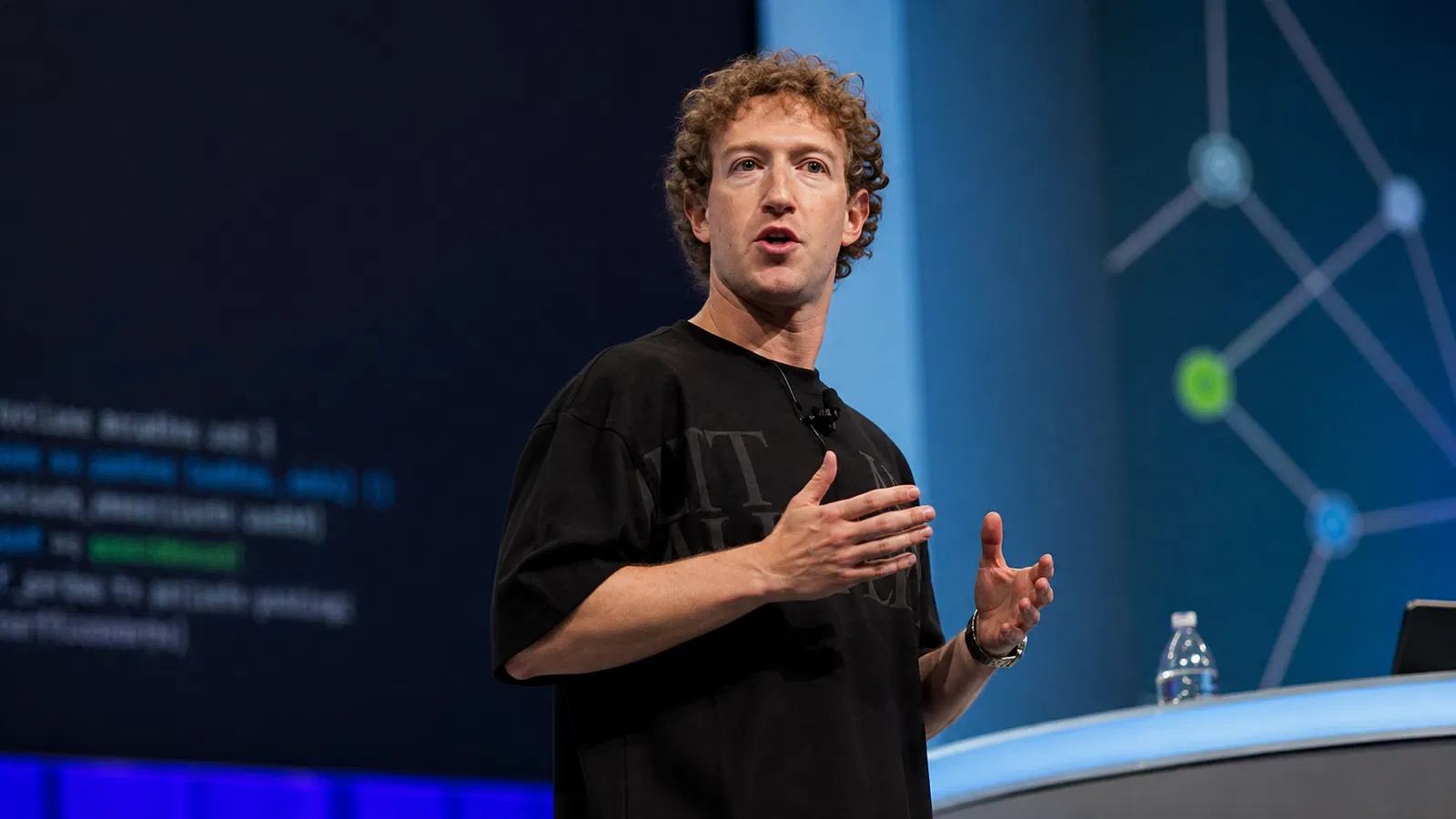 Foto real de Mark Zuckerberg em palco, com identidade Meta discreta e público ao fundo.